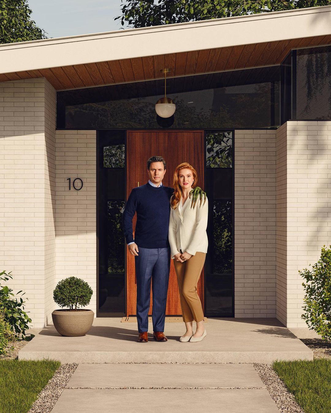 A man and woman stand together on the porch of a modern home with white brick and a brown door, a small tree in a pot nearby.