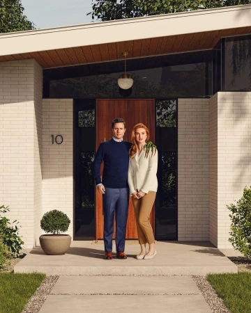 A man and woman stand together on the porch of a modern home with white brick and a brown door, a small tree in a pot nearby.