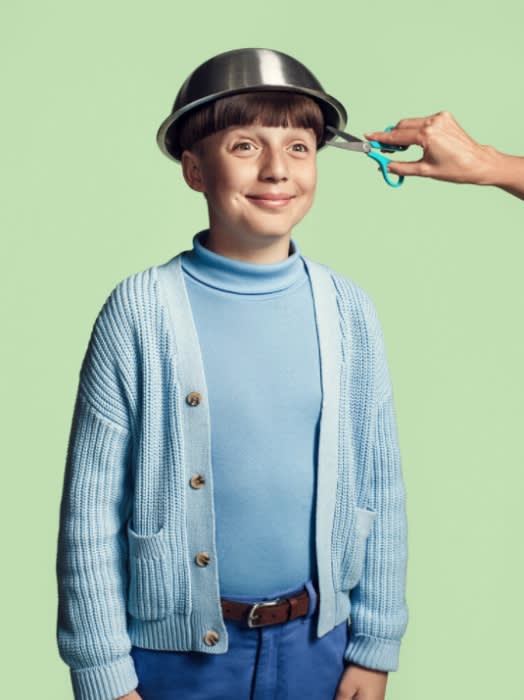 Boy getting a haircut with a metal bowl on his head, light blue sweater, blue pants, and a green background. 