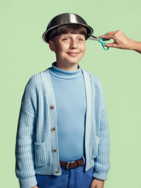 Boy getting a haircut with a metal bowl on his head, light blue sweater, blue pants, and a green background. 