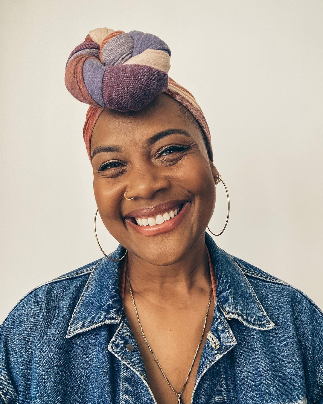 Smiling woman with dark skin and a colorful head wrap, wearing a denim jacket, looking directly at the camera, against a plain background.