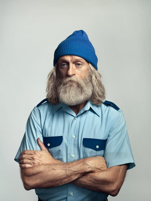 Portrait of an adult male with a long beard wearing a blue knit hat and a light blue short sleeve button down shirt with arm crossed.