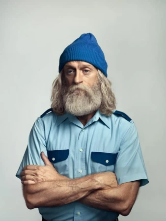 Portrait of an adult male with a long beard wearing a blue knit hat and a light blue short sleeve button down shirt with arm crossed.