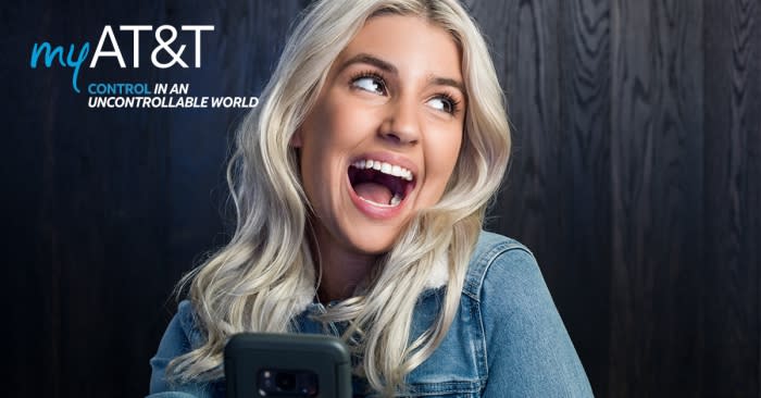 Smiling woman with platinum blonde hair in a denim jacket looks up while holding a smartphone against a grey wood plank background.