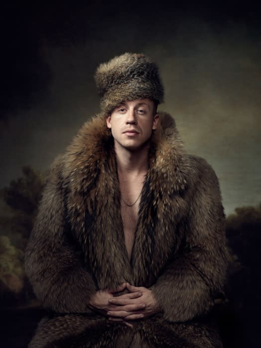 Man in fur hat and coat with clasped hands in studio portrait with a dark, painterly background, wearing gold chain, neutral expression.
