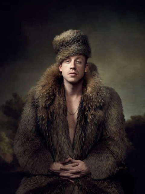 Man in fur hat and coat with clasped hands in studio portrait with a dark, painterly background, wearing gold chain, neutral expression.