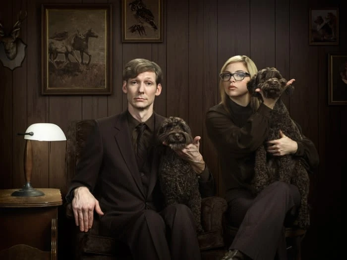 A studio portrait captures a man and woman holding brown Poodle dogs while sitting on a couch in front of wood-paneled walls.