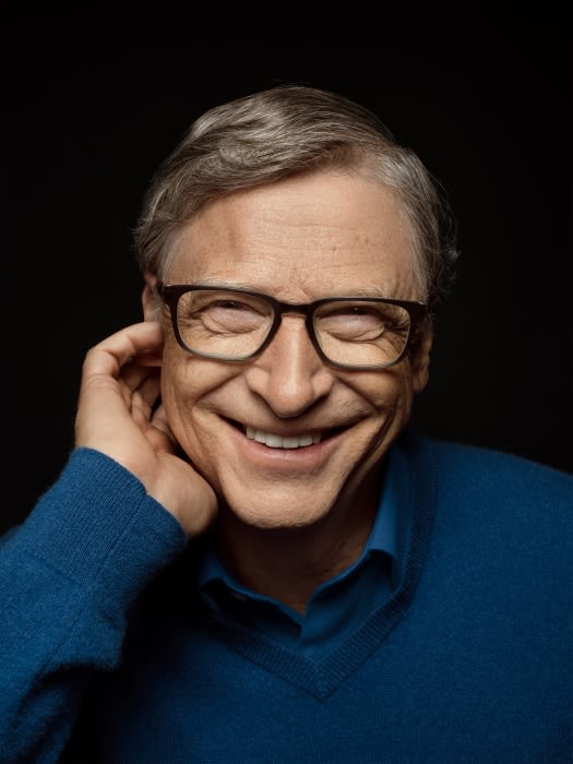Portrait of an adult man with gray hair wearing glasses and a blue sweater, smiling with hand near face against a dark background.