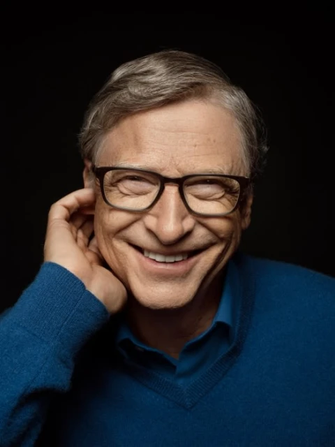 Portrait of an adult man with gray hair wearing glasses and a blue sweater, smiling with hand near face against a dark background.