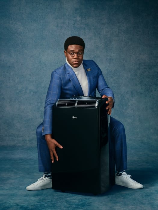 Adult male in a blue suit posing with a black portable air conditioner against a mottled blue backdrop in a studio setting.