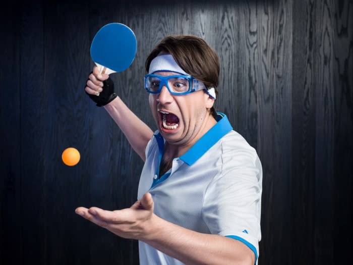 Enthusiastic man in eyewear with table tennis paddle and ball against a wood-paneled background, expressing excitement and focus.