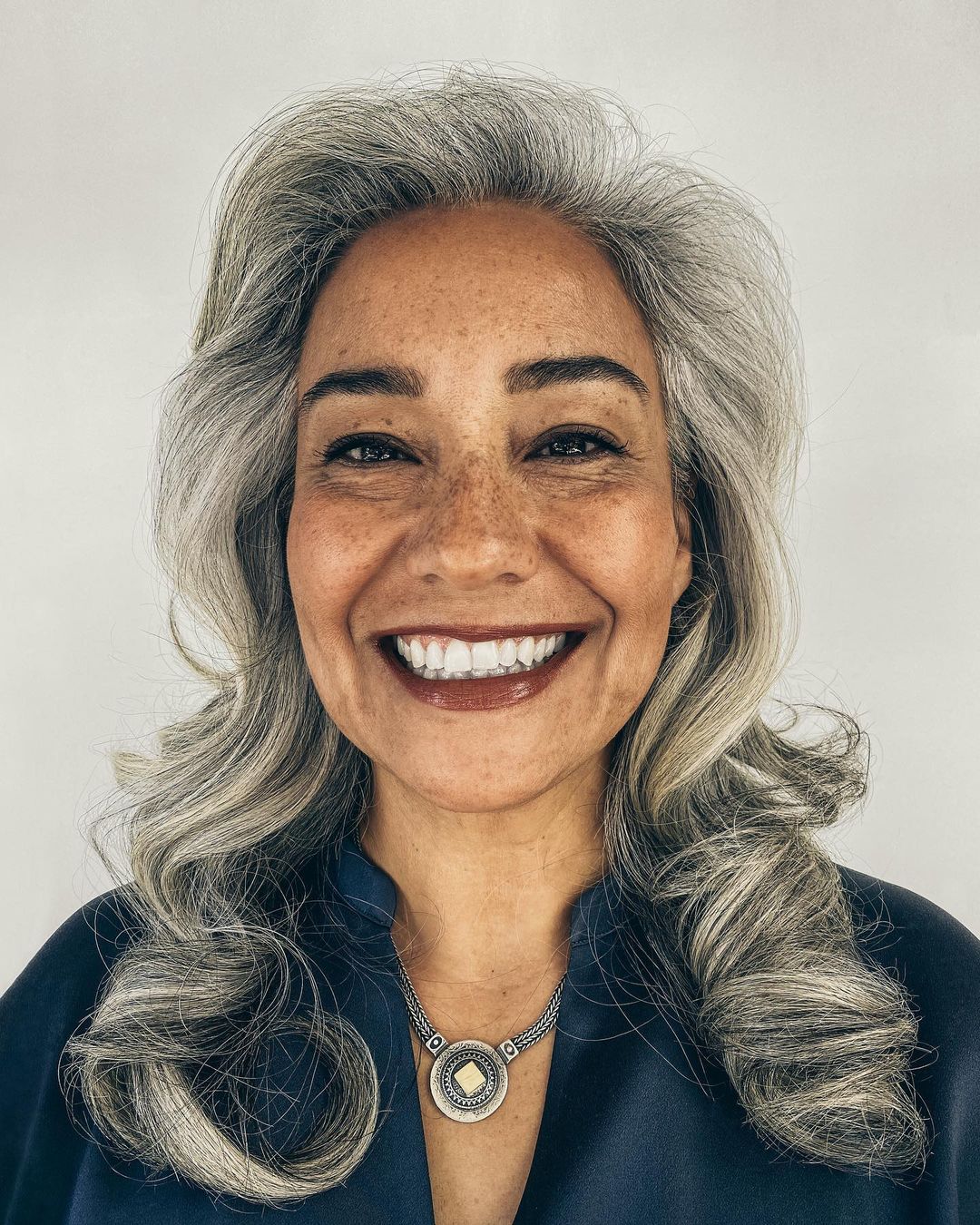 Close-up portrait of a mature adult female with gray hair smiling, wearing a blue top and silver pendant against a white background.