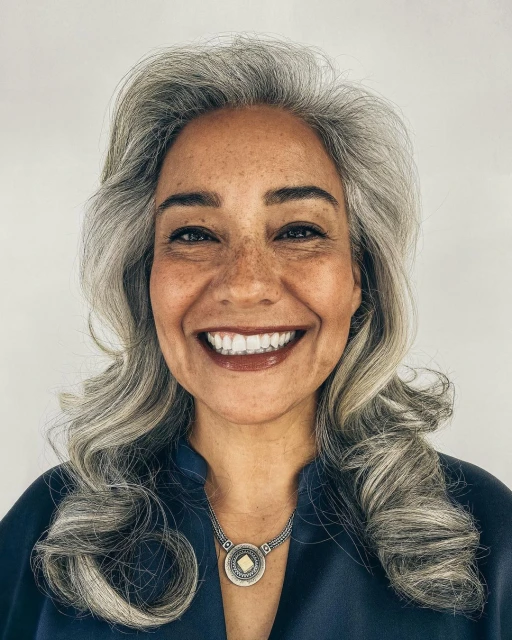 Close-up portrait of a mature adult female with gray hair smiling, wearing a blue top and silver pendant against a white background.