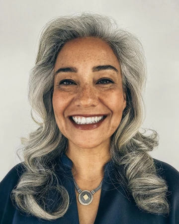 Close-up portrait of a mature adult female with gray hair smiling, wearing a blue top and silver pendant against a white background.