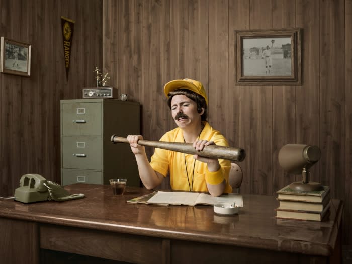Humorous portrait of a man dressed as a vintage baseball coach sitting at a desk with a baseball bat and vintage office equipment.