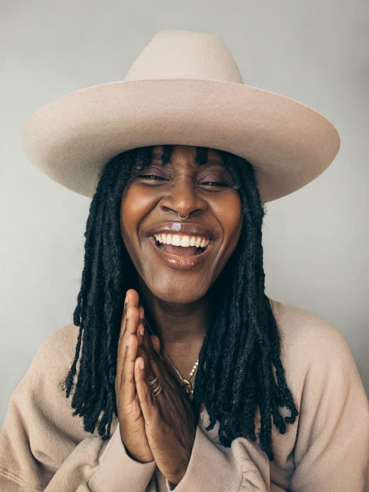 Portrait of a smiling adult female wearing a beige hat and sweater with hands clasped. Neutral background, indoor setting, medium shot.