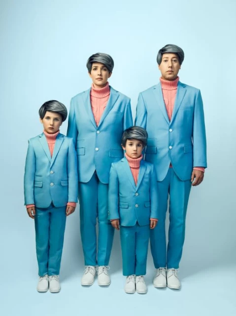 A family of four poses in matching light blue suits and pink turtlenecks, set against a light blue backdrop in a portrait format.