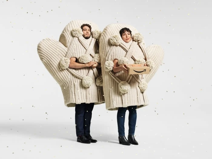 Two people wearing knit mitten costumes holding an Amazon box in a bright white studio setting with gold confetti accents.