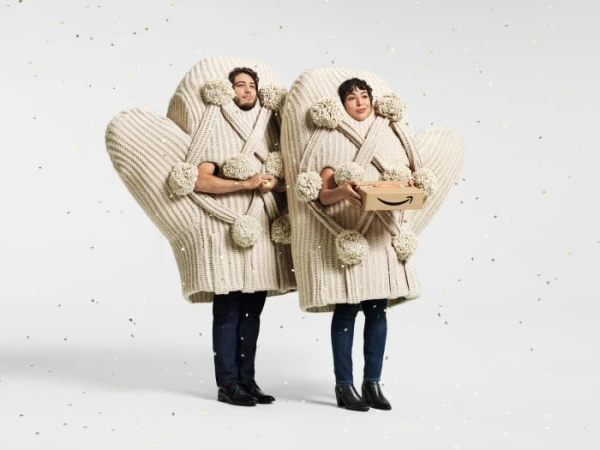 Two people wearing knit mitten costumes holding an Amazon box in a bright white studio setting with gold confetti accents.