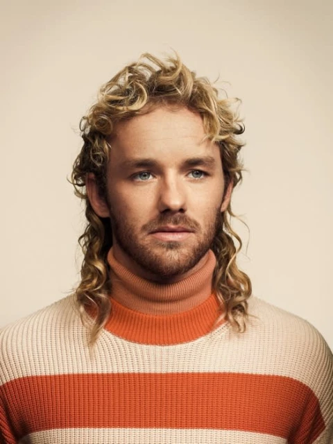 Portrait of a young adult male with blond curly hair wearing a cream and orange striped turtleneck sweater against a neutral background.