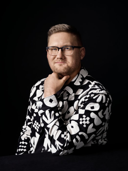 Portrait of an adult male with short brown hair, glasses, and a black and white patterned jacket posing against a dark backdrop.
