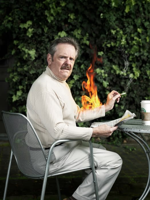 Mature man in a white turtleneck, seated at a metal table outdoors, reading a newspaper with an orange flame superimposed on his arm.