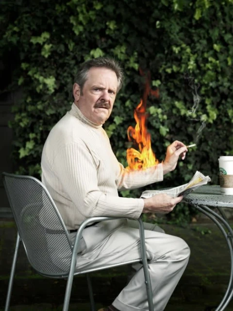 Mature man in a white turtleneck, seated at a metal table outdoors, reading a newspaper with an orange flame superimposed on his arm.