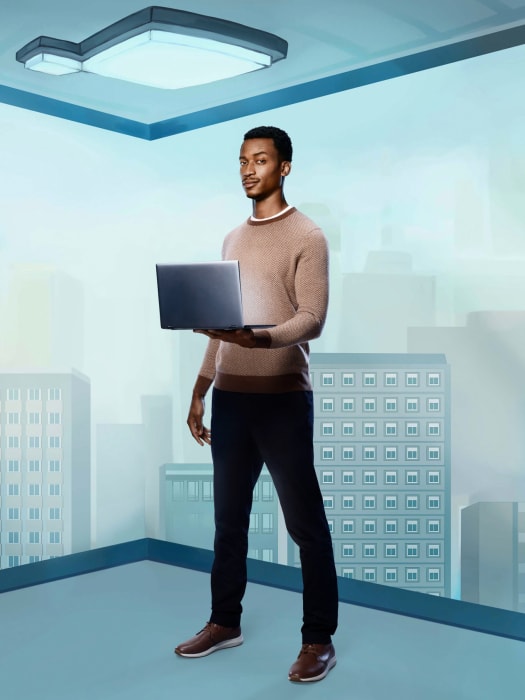 An African-American man holding a laptop stands indoors, wearing casual clothes, in an environment with blue walls and large windows.
