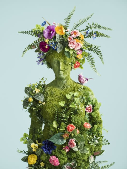 Moss-covered statue adorned with flowers and greenery, bird perched on shoulder, against a light blue background. Lush and botanical.