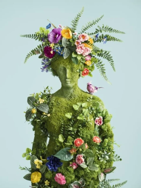 Moss-covered statue adorned with flowers and greenery, bird perched on shoulder, against a light blue background. Lush and botanical.