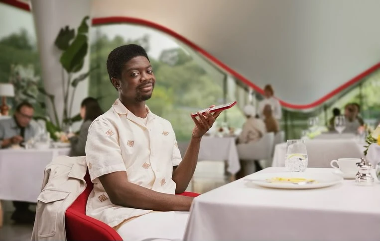 African American man in a patterned shirt sits in a restaurant holding a smartphone, other diners and plants visible in soft focus.