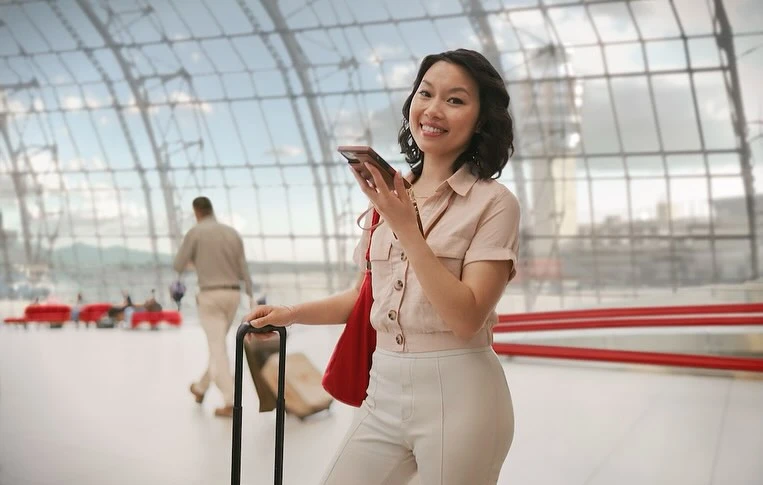 Smiling woman in airport using smartphone, pulling rolling suitcase with red shoulder bag. Large windows, modern architecture.