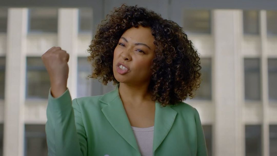 Adult female wearing a green jacket raises her fist in an indoor environment with natural light in front of an office building background.