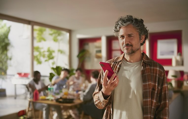 Adult male holds a phone in an indoor gathering with a group of people at a table, illuminated by indoor lighting and natural daylight.