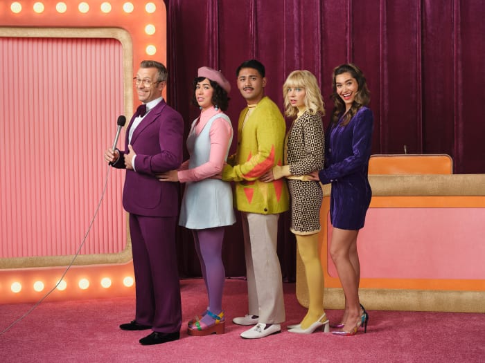 Five people stand in a line on a pink stage for a game show. A host in a purple suit holds a microphone on the left.