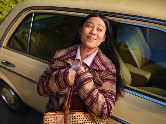 Smiling Asian woman with handbag stands by vintage car. She's wearing a patterned jacket and light purple shirt in daylight conditions.
