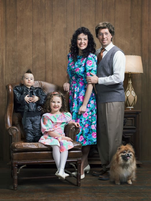 Family portrait in 1980s style features parents, two children, and a dog indoors with wood-paneled walls and vintage furniture and clothing.