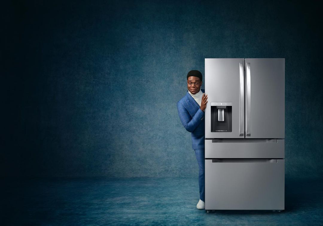 An African American man wearing a blue suit peeks from behind a modern, stainless steel refrigerator in a cool-toned studio setting.
