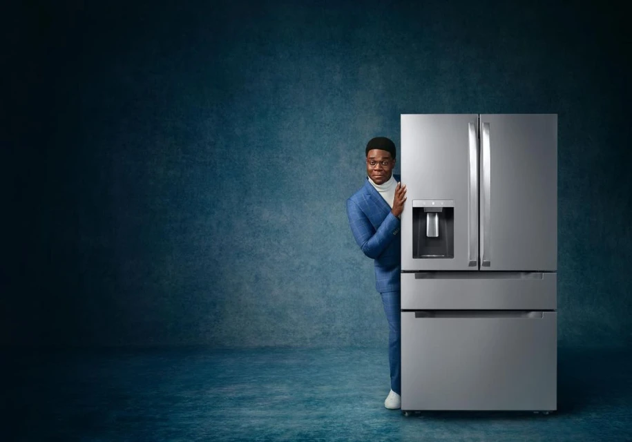 An African American man wearing a blue suit peeks from behind a modern, stainless steel refrigerator in a cool-toned studio setting.