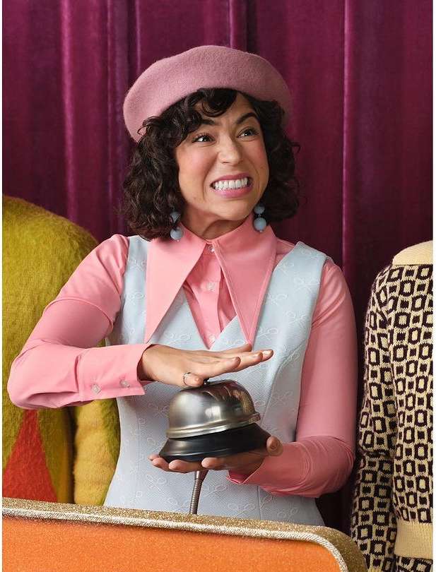 Adult female ringing a silver service bell while wearing a pink beret, light pink top and light blue vest against a draped maroon background.