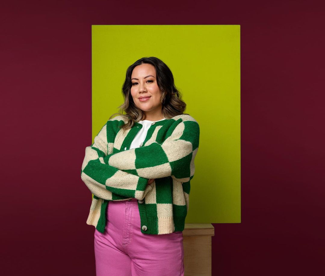 Portrait of an adult woman with crossed arms, wearing a green and white checkered cardigan and pink pants, standing in front of a green backdrop.