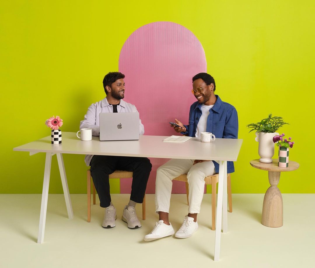 Two adult men sit at a white desk with a laptop and smartphone, smiling in a brightly lit studio with a pink and yellow background.