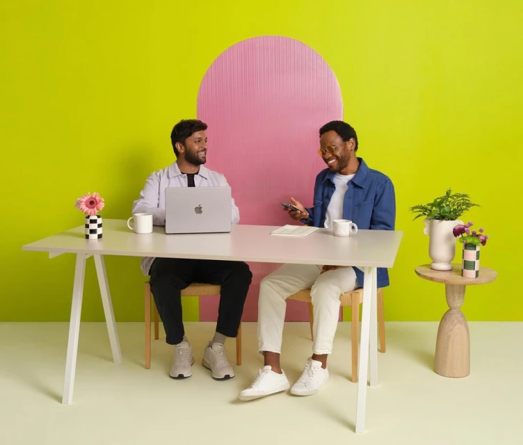 Two adult men sit at a white desk with a laptop and smartphone, smiling in a brightly lit studio with a pink and yellow background.