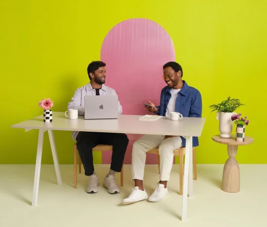 Two adult men sit at a white desk with a laptop and smartphone, smiling in a brightly lit studio with a pink and yellow background.