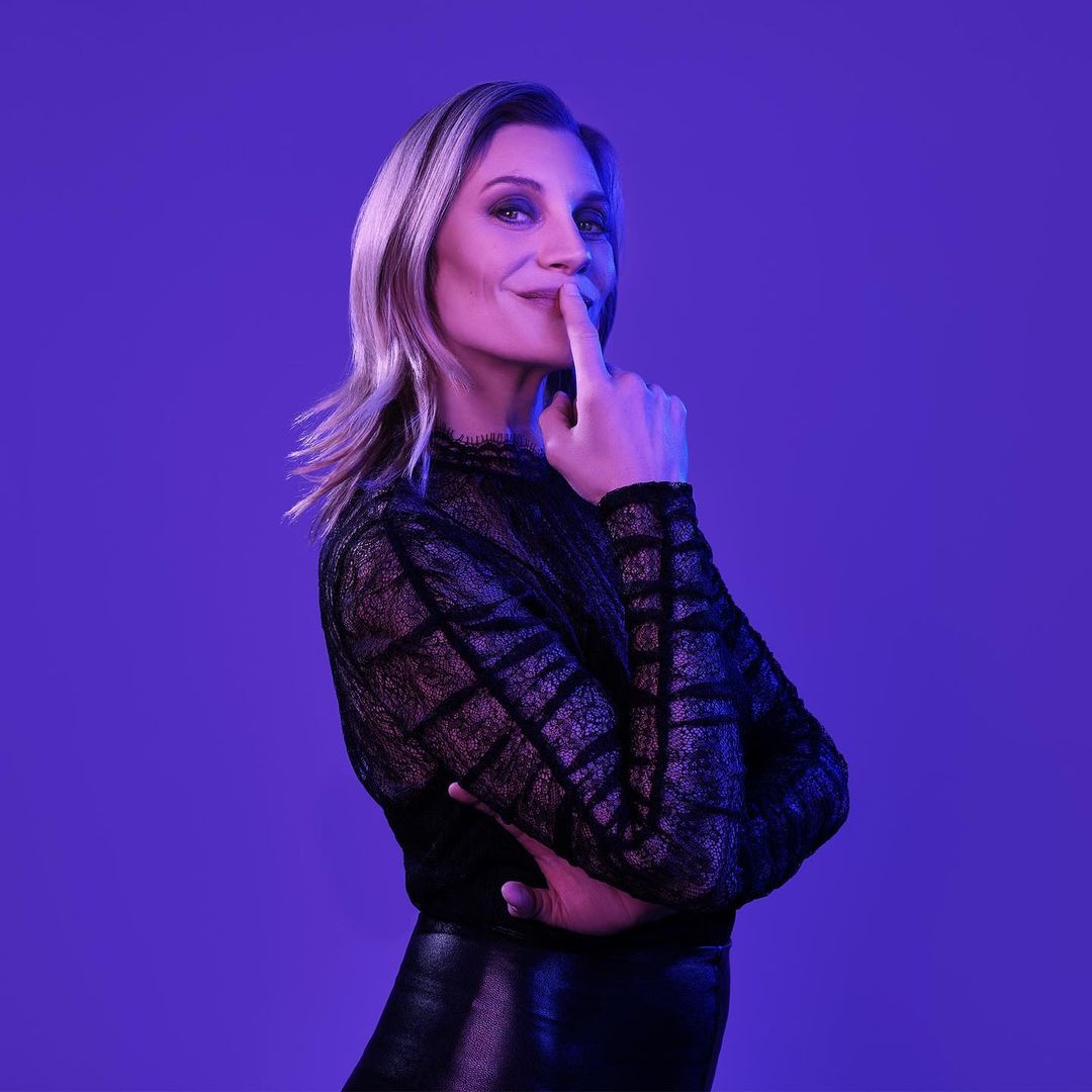 A woman with blonde hair poses with her finger to her lips in front of a solid purple background, wearing a black lace top.