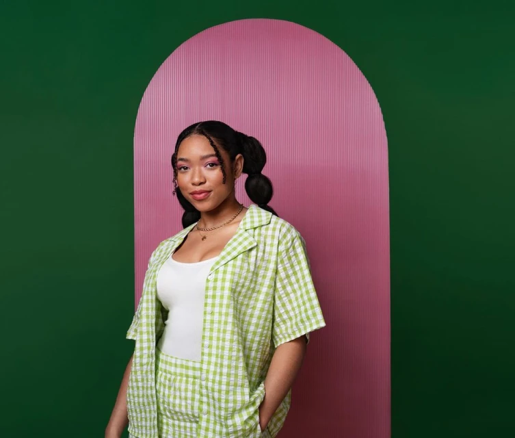 Young woman in a green and white checkered outfit poses in front of a green and pink background with a rounded doorway shape.