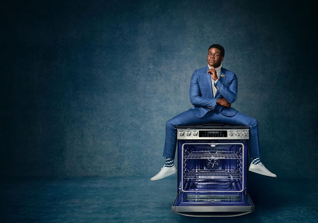 Adult male in suit sitting on oven in studio, wearing sneakers, looking off to the side, against a mottled blue background.
