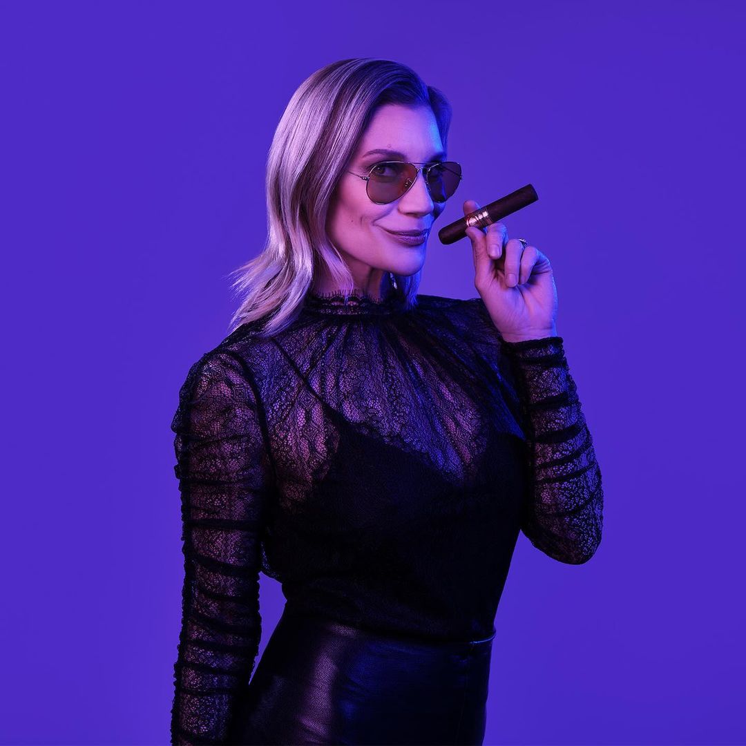 Blonde woman with sunglasses holds cigar in a studio lit with blue and purple tones, wearing a black lace top and leather skirt.