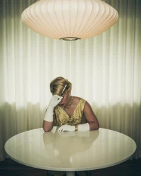 Woman in vintage yellow dress and white gloves seated at a round table under a large pendant light