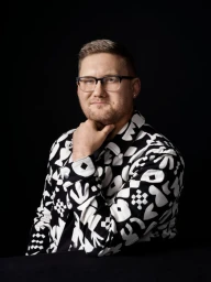 A man wearing glasses and a patterned black and white shirt posing against a solid black background.